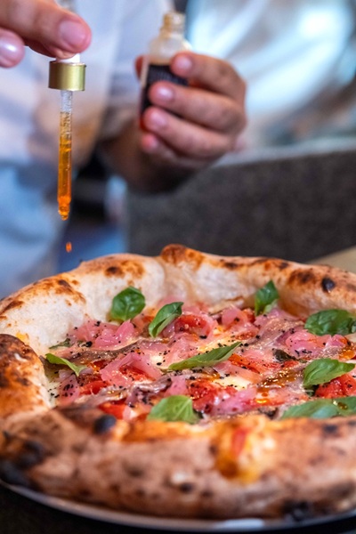 Pizza dal cornicione bruciacchiato con prosciutto, pomodorini e basilico, mentre un contagocce versa olio arancione